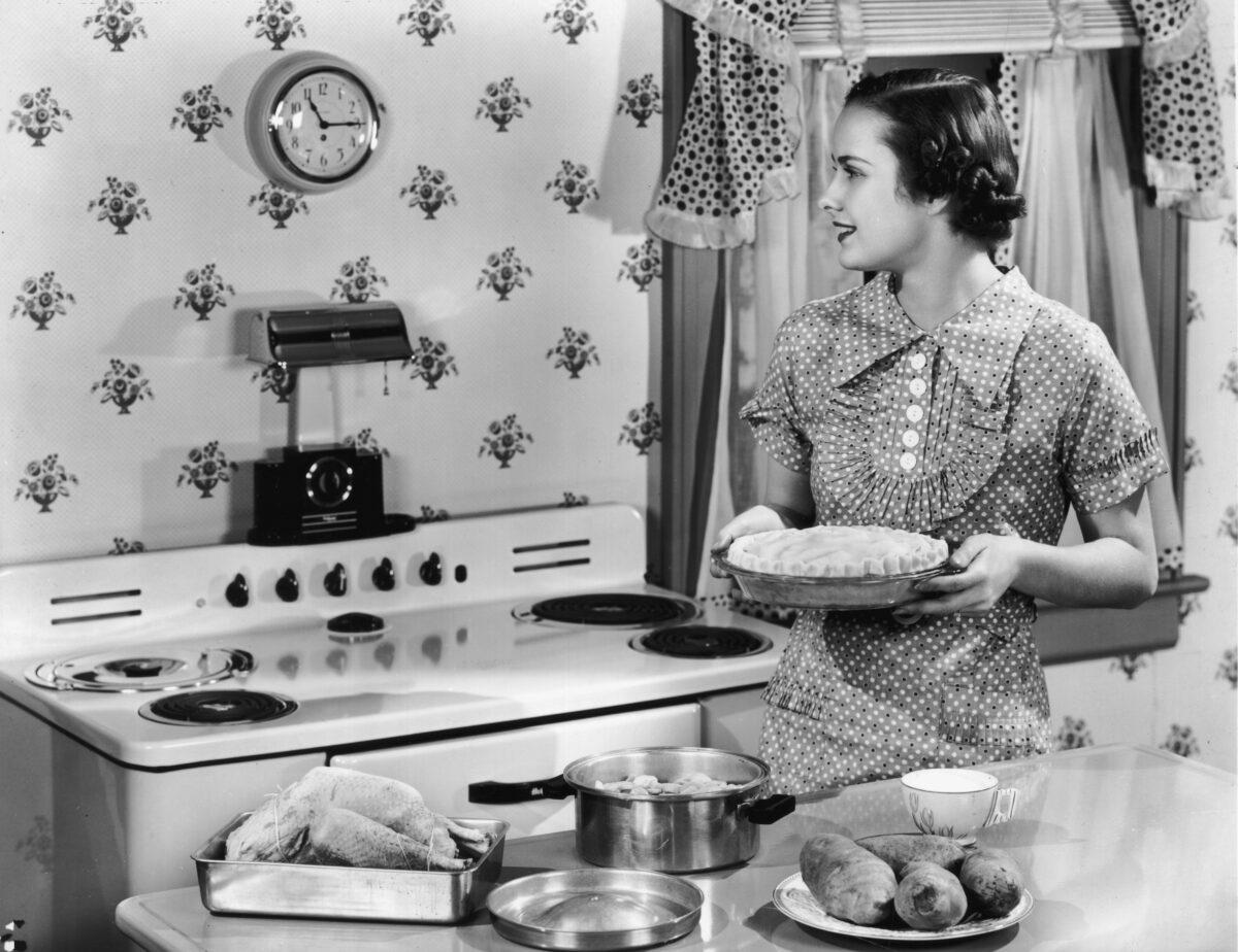 A housewife holds an unbaked pie while looking at a wall clock in a kitchen. An unprepared roast chicken and potatoes sit on the counter. (Hulton Archive/Getty Images)