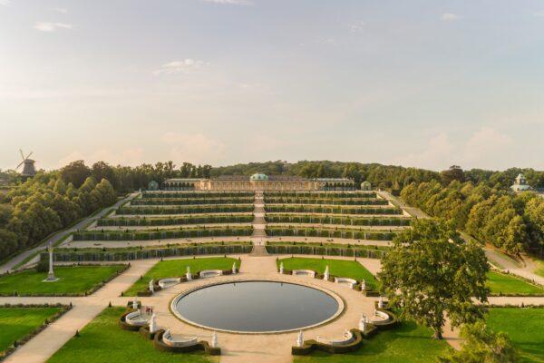 An aerial view of the Sanssouci complex. The world-renowned park features terraced grounds and a majestic fountain at the center, surrounded by marble benches and sculptures representing Olympian gods. The baroque parterre near the palace features more than 230,000 plants. (With permission, © SPSG/Reinhardt und Sommer)
