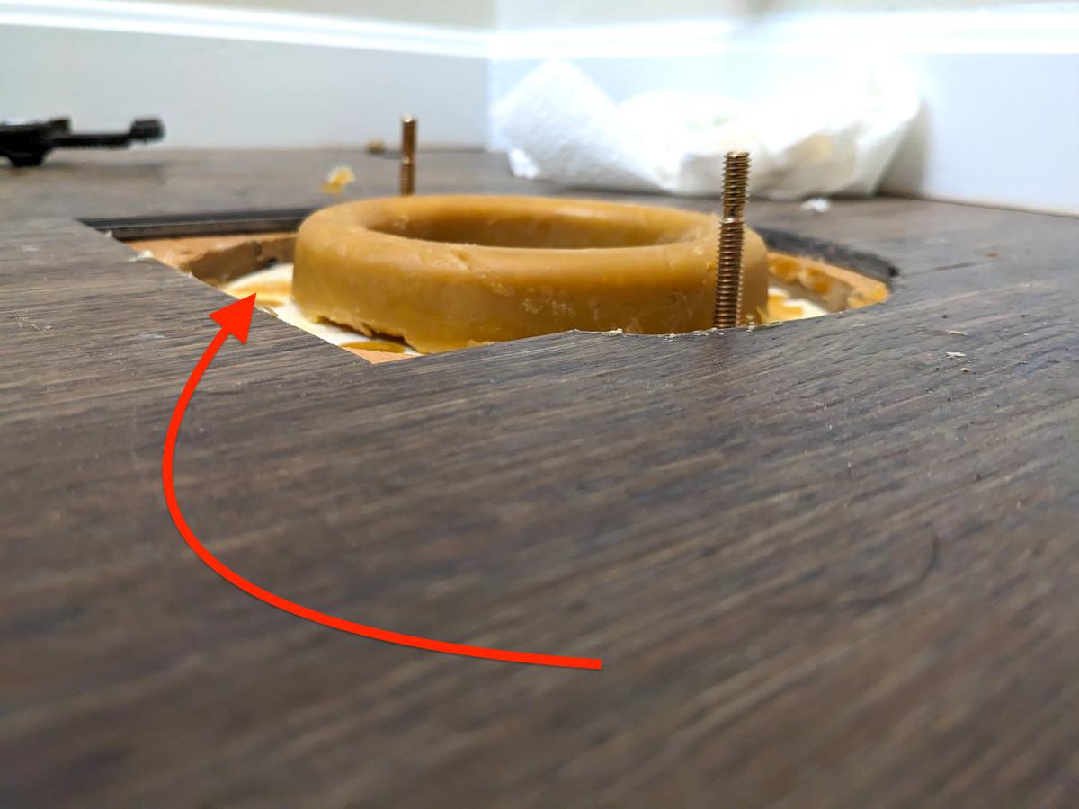 The red arrow points to the white toilet flange that’s currently buried under two layers of laminate flooring. It was supposed to be installed on top of the flooring! (Tim Carter)