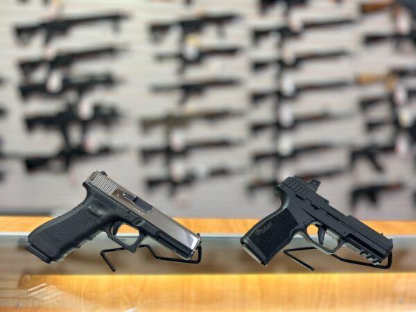 A Glock 17 9 mm (L) and a Sig Sauer P322 .22-caliber handgun are propped up by stands on a glass countertop in front of a wall of rifles in Lawful Defense in Gainesville, Fla., on April 19, 2023. (Nanette Holt/The Epoch Times)