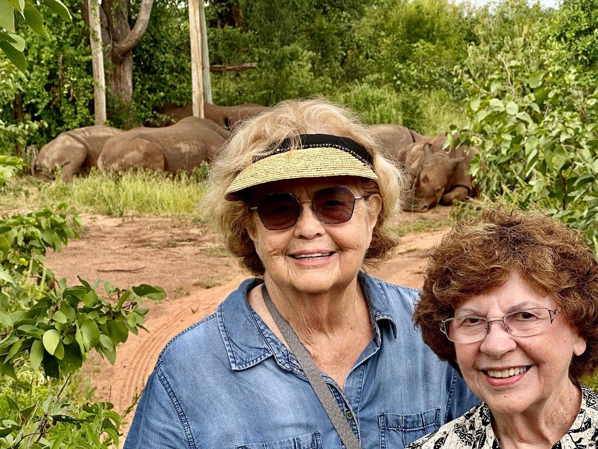 Hazelip and Ellie pose for a photo as five white rhinos, in the background, rest in the shade during the hot afternoon sun in Zambia. (Courtesy of Around the World at 80)