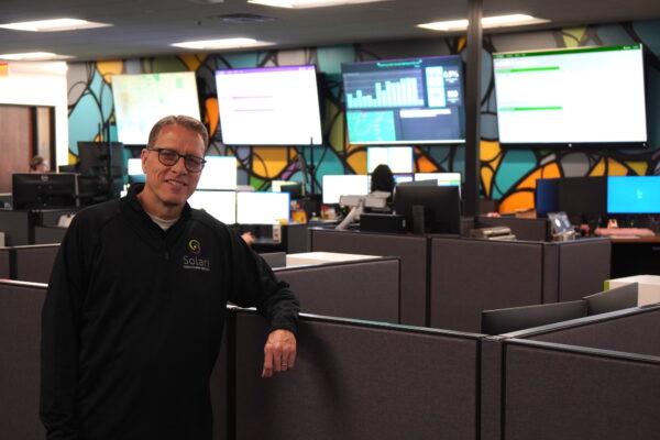 Thomas Bond, senior director of communications, marketing, and education at Solari Crisis and Human Services in Tempe, Ariz., stands in front of a complex array of computer monitors on April 13, 2023. (Allan Stein/The Epoch Times)