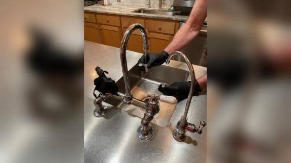 A USGS scientist wearing black gloves is collecting a sample of tap water from the kitchen sink using small plastic vials to test for PFAS. (Paul Bradley/USGS)