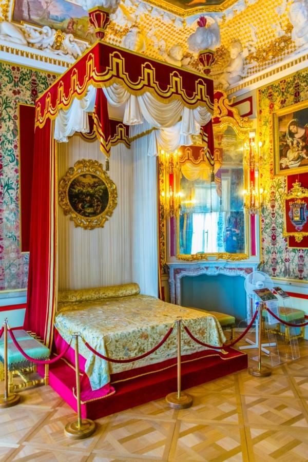 Genoa-style velvet patterned wallpaper lines the walls of the queen’s bedroom. A reconstructed bed, based on a design by Daniel Marot (a draftsman who served King Louis XIV), features a red and gold canopy topped with two feather plumes. Putti and sphinxes feature throughout the ceiling stucco work, along with springtime frescoes quoting and illustrating Virgil’s “Georgics.” Spring motifs also run around the large Regency mirror frames, reflecting the opulent room. (<a href="https://www.shutterstock.com/image-photo/warsaw-poland-august-13-2016-view-642288961">trabantos</a>/Shutterstock)
