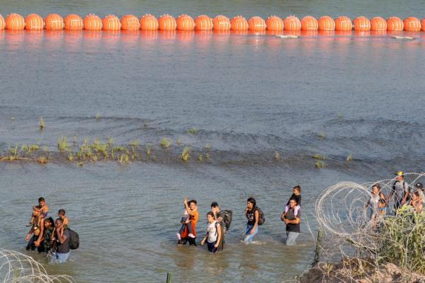People walk between razor wire and a string of buoys placed on the water along the Rio Grande border with Mexico in Eagle Pass, Texas, on July 16, 2023. (Suzanne Cordeiro/AFP/Getty Images)