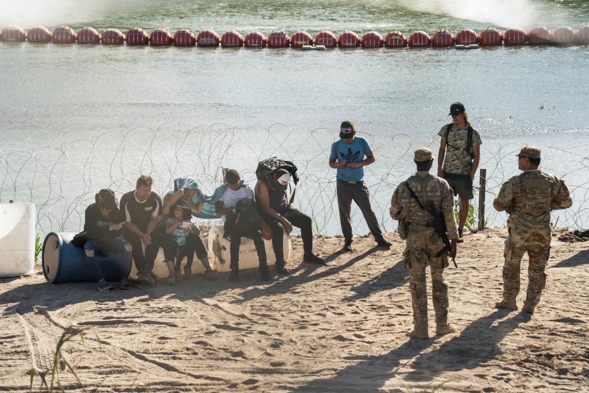 Asylum-seeking illegal immigrants sit by a concertina wire fence while waiting to be transported by U.S. law enforcement officers after crossing the Rio Grande river into the U.S. from Mexico in Eagle Pass, Texas, on July 24, 2023. (Go Nakamura/Reuters)