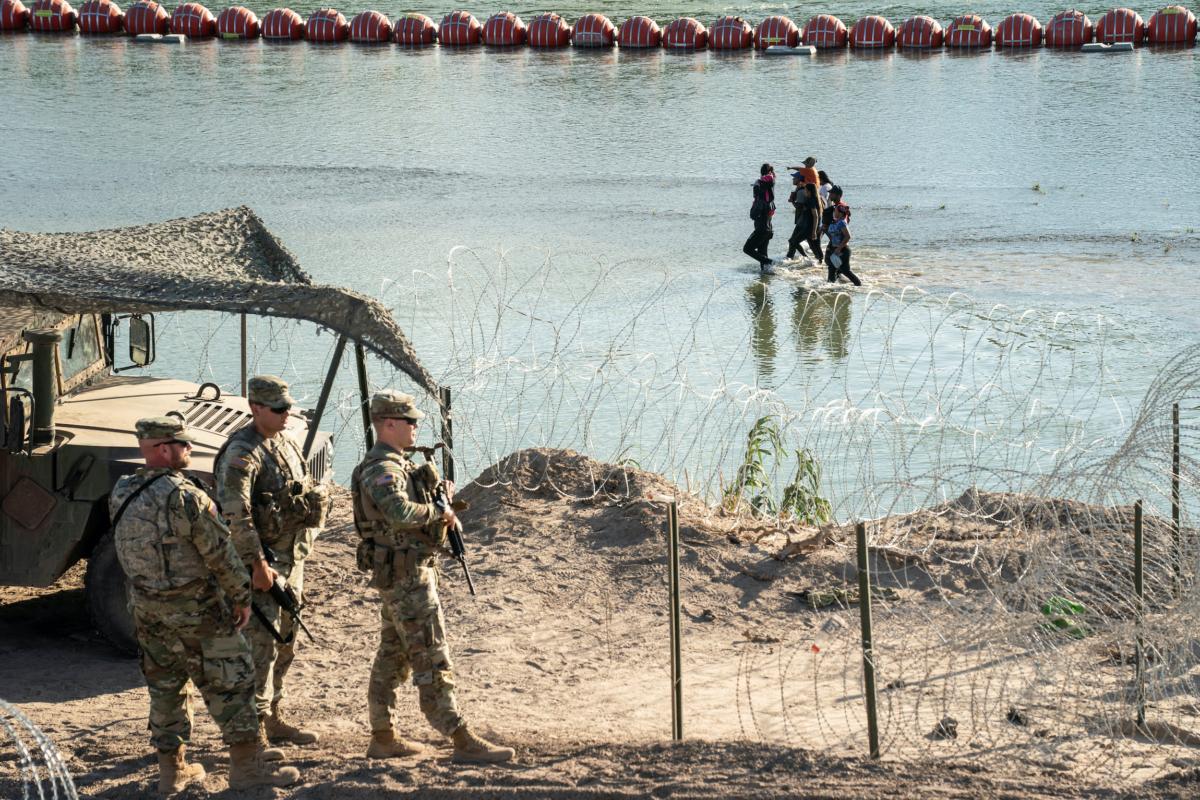 Illegal immigrants walk in the Rio Grande river between a floating fence and the river bank as they look for an opening on the concertina wire fence to land on U.S. soil in Eagle Pass, Texas, on July 24, 2023. (Go Nakamura/Reuters)