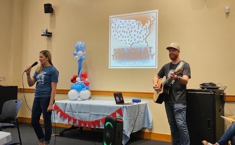 Sarah Fenske (L) leads the group in songs, accompanied by a father playing his guitar at the Rohnert Park Library during the Sonoma County story hour in Rohnert Park, Calif., on Aug. 5, 2023. A slide is shown on the wall of some of the places across the nation where Brave Books story hours were being presented on Aug. 5. (Courtesy of Sarah Fenske)