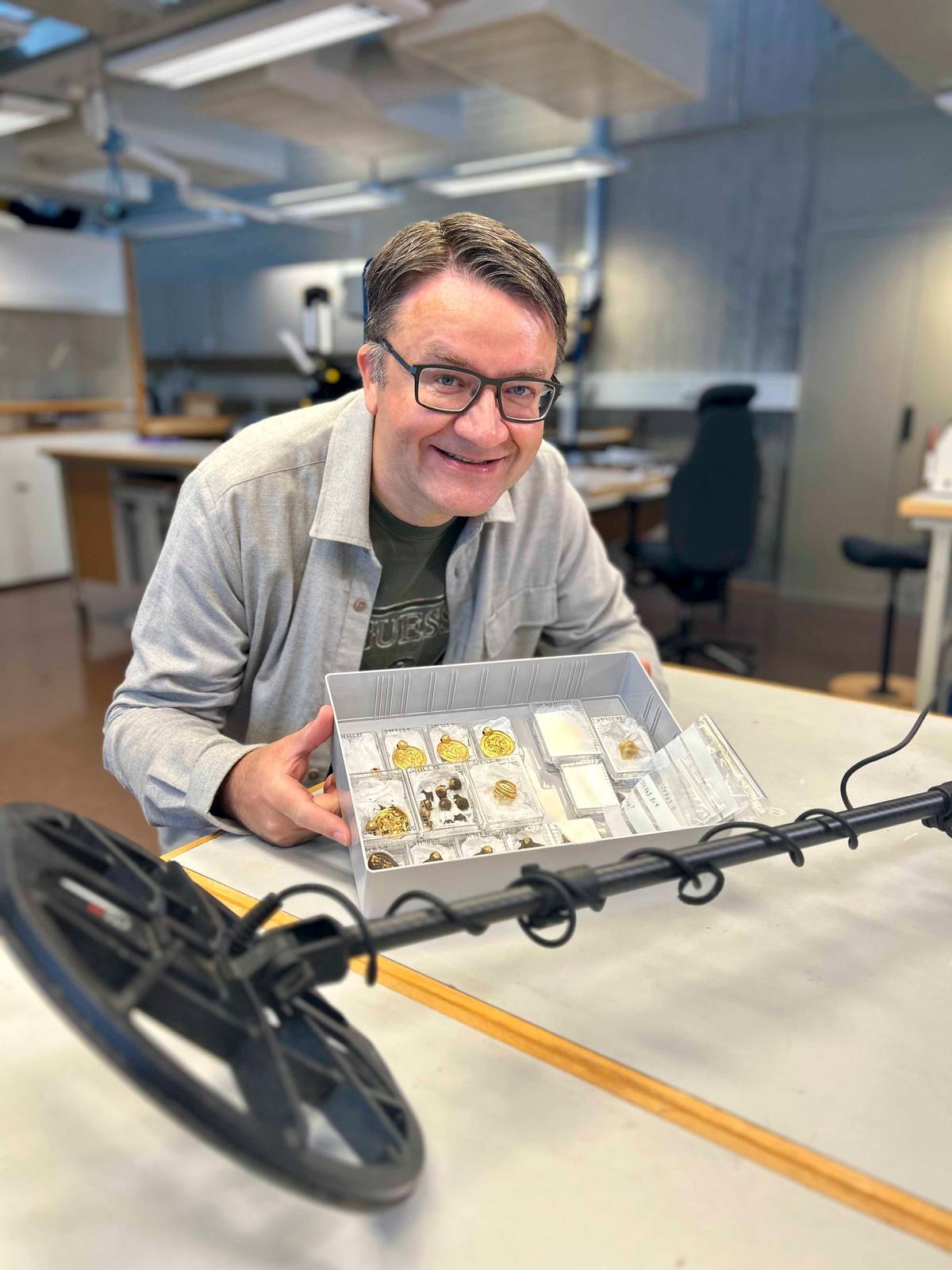 Mr. Bore with the 6th-century gold jewelry and his metal detector. Initially, he thought these were some "chocolate coins or plastic pirate treasure." (© Anniken Celine Berger – The Museum of Archaeology, University of Stavanger)