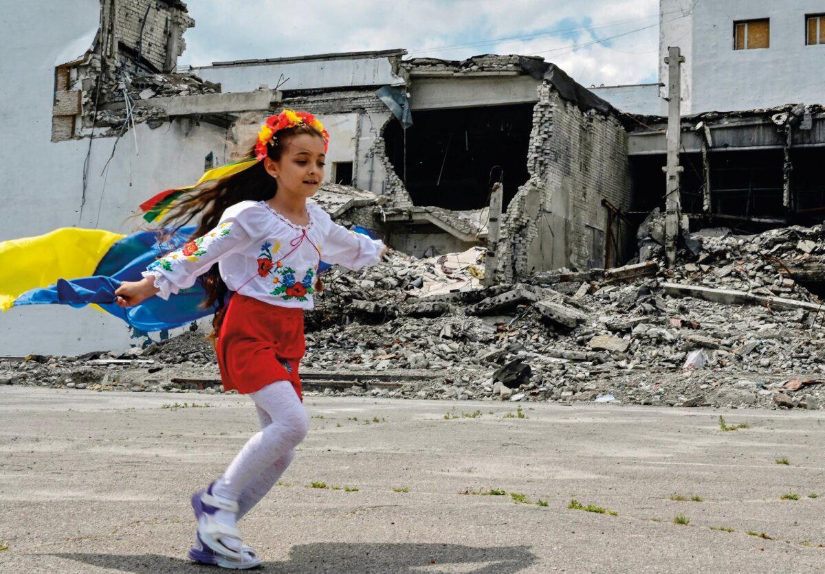 A young girl holding a Ukrainian flag runs in front of a destroyed cultural center during the graduation ceremony of art students in the town of Derhachi in the Kharkiv region of eastern Ukraine. (Sergey Bobok/Contributor/AFP/ Getty Images)