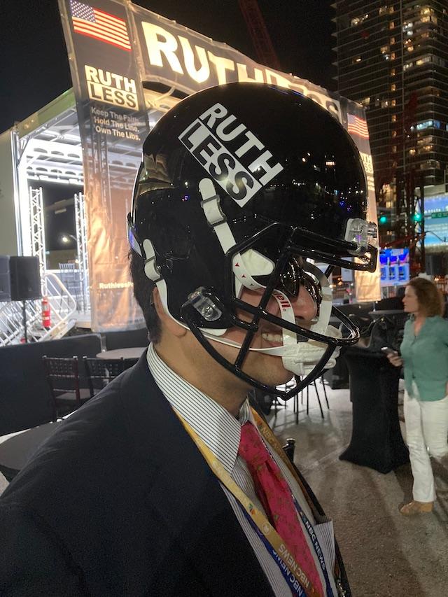 Ruthless Podcast's Comfortably Smug, who says everyone knows his name, can't fit a football helmet on his head during a parking lot GOP debate watch party on Nov. 8, one of few people to be found anywhere near the venue as five candidates took the stage. (John Haughey/The Epoch Times)