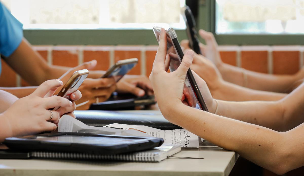 High school students sit at a table holding multiple digital devices such as phones and tablets during a class lesson. (LBeddoe/Shutterstock)