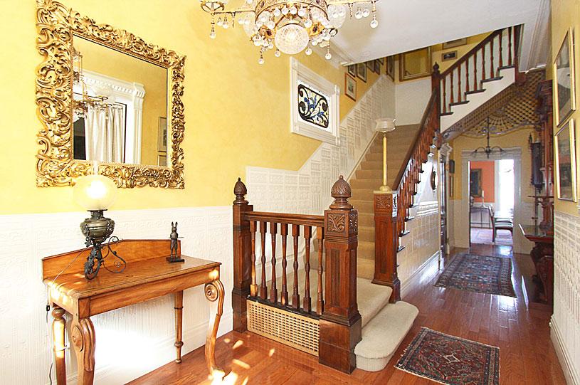 The foyer and front staircase of 722 Steiner Street were maintained in the Victorian style until a renovation in 2022. The chandelier is most likely original, as are the gilded mirror and ornately carved wooden posts on the stairs. The detailed wood carving under the staircase was completed by the original builders. All floors are wood, highly polished, and covered with Persian carpets. (Courtesy of Linnea Key Realty)