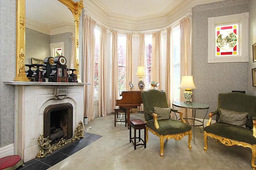 This room, shown here before the 2022 renovation, hosted a baby grand piano in the bay window, presumably for all to see.  The fireplace was topped with an ornate gilded mirror with brass fixtures on the tiled hearth. Richly upholstered furniture with gilded trim was the perfect way to seat visitors for a piano recital. The stained-glass window and the accessories on top of the fireplace showed the wealth of the owner. (Courtesy of Linnea Key Realty)
