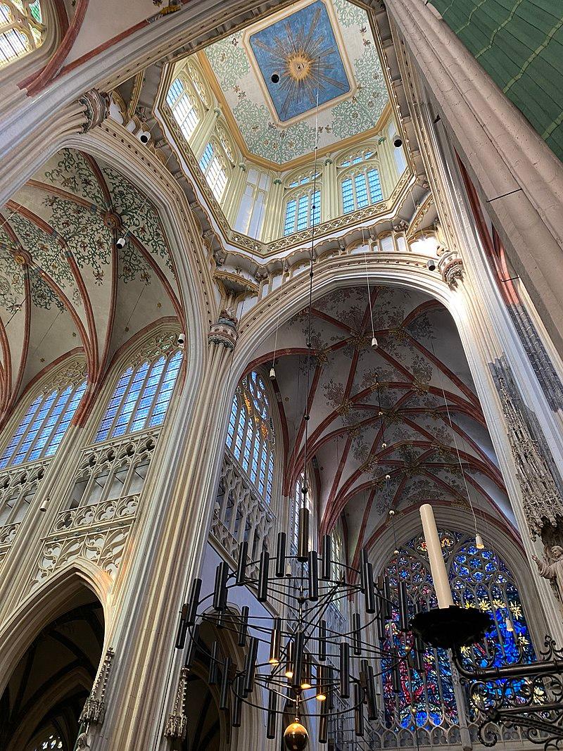 The cathedral's vaulted ceiling is supported by tall slender columns. The light-colored ceiling features a delicate and colorful sun across a blue background surrounded by intricate leaves and foliage. The pointed arched windows and stainless-steel glass allows in natural light. (Maarten1703/CC BY-SA 4.0)