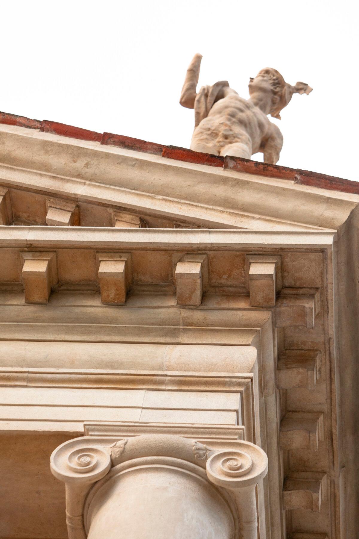 The finely carved Ionic stone scrolls have a gentle quality and are emphasized when juxtaposed with the otherwise plain entablature (horizontal band above the columns) and the repetition of corbels (rectangular boxes) above. A classical sculpture stands up top. (J.H. Smith)