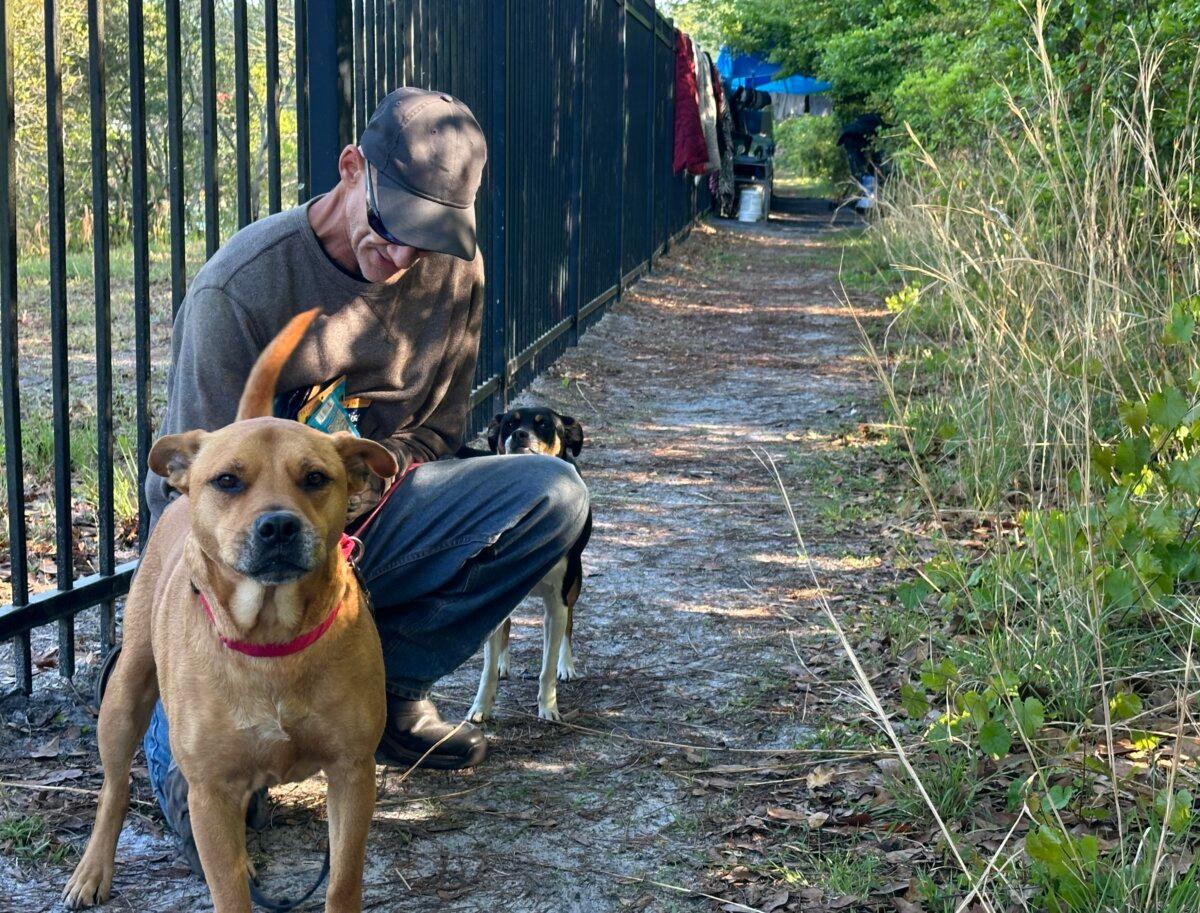 Danny, who asked to be identified by his first name only, sits outside his camp near a shopping center in Gainesville, Fla., with his dogs Brody and Bella on March 20, 2024. (Nanette Holt/The Epoch Times)