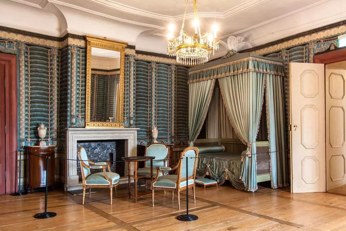 Located on the second floor, the Von Hochberg Apartment, named after the countess who resided there, is furnished in the 19th-century Empire style. The bedroom includes walnut chairs with green upholstery, a canopy bed, a fireplace, and classic vases. (Courtesy of <a href="https://www.schloss-schwetzingen.de/fileadmin/_processed_/8/f/csm_37_schwetzingen_innen_app-luise-von_hochberg_wohnzimmer_foto-ssg-ursula-wetzel_569dc021fb.webp 7 https://commons.wikimedia.org/wiki/File:Schwetzingen-n%C3%B6rdlicherZirkelbau.jpg" target="_blank" rel="nofollow noopener">State Palaces and Gardens of Baden-Württemberg</a>)