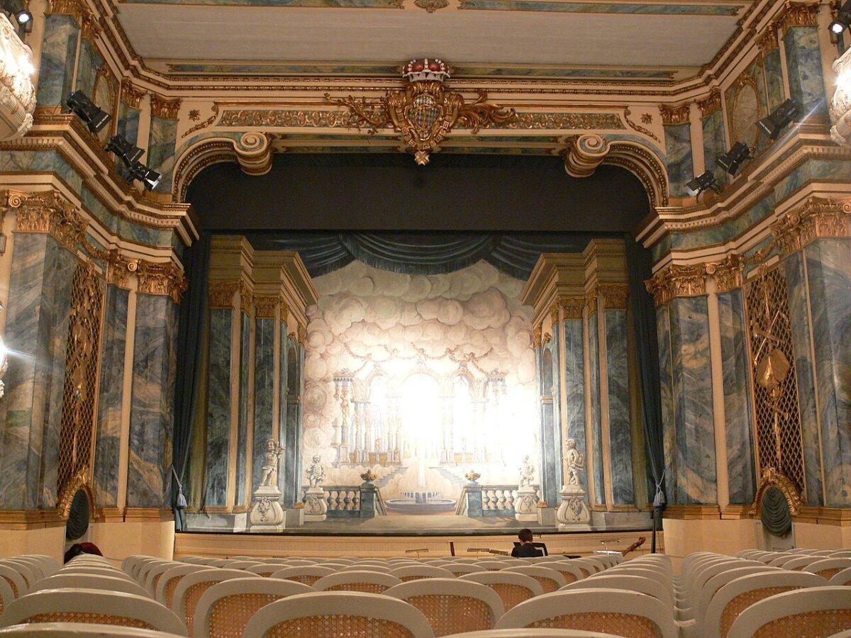 The Schwetzingen theater contains a unique blend of the rococo and neoclassical styles with its gilded features and stucco work. The center stage is framed by marble columns and displays an elaborate baroque set with complex stage machinery. On both sides, elegant stucco galleries feature tiers without boxes, inspired by French and Italian court theaters of the day. (<a href="https://en.wikipedia.org/wiki/Schwetzingen_Palace#/media/File:Schwetzingen_Schlosstheater_Blick_zur_B%C3%BChne_1.jpg" target="_blank" rel="nofollow noopener">Andreas Praefcke/CC BY 3.0</a>)