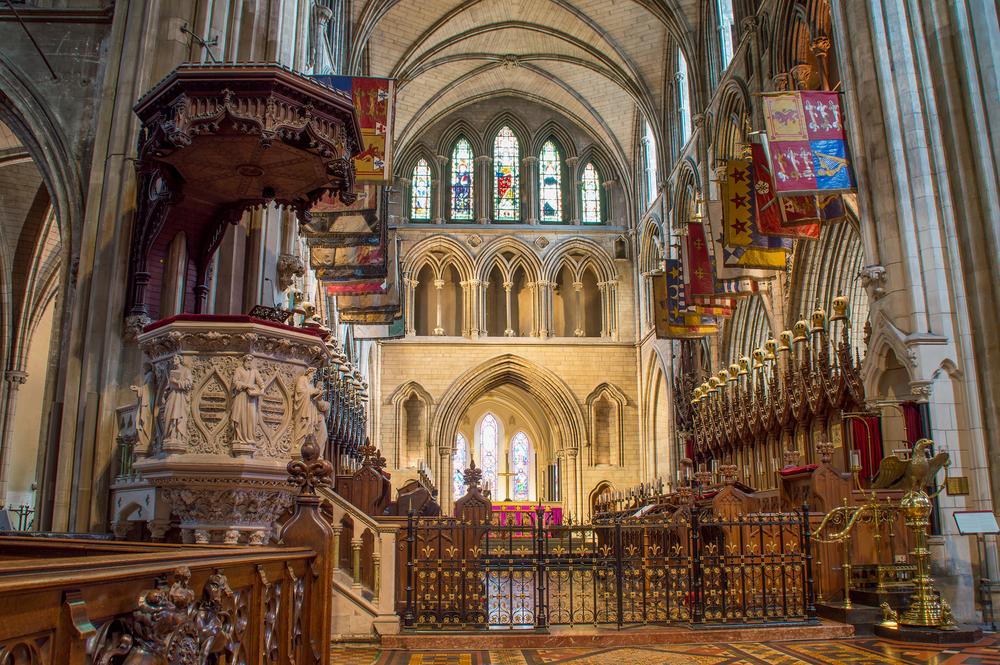 The pulpit on the left, with intricate wood carvings and detail, stands as a focal point within the cathedral. Next to the pulpit, the choir is framed by symmetrical pointed arches and a vaulted ceiling. At the heart of the choir lies the sanctuary, featuring the high altar and stained-glass windows with images of Christ at the center, surrounded by the four evangelists: Matthew, Mark, Luke, and John. (Thoom/Shutterstock)