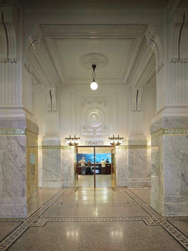 Fluted columns, decorative moldings, and a medallion from which a simple brass-fitted globe light hangs greet visitors and customers in the ticket hall. This hall’s design shows the glittering influence of the Gilded Age (late 1800s to early 1900s), with marble column bases accentuated by a gilded band and polished brass doors that lead to the Amtrak ticket desk. (Courtesy Photographer Benjamin Benschneider)