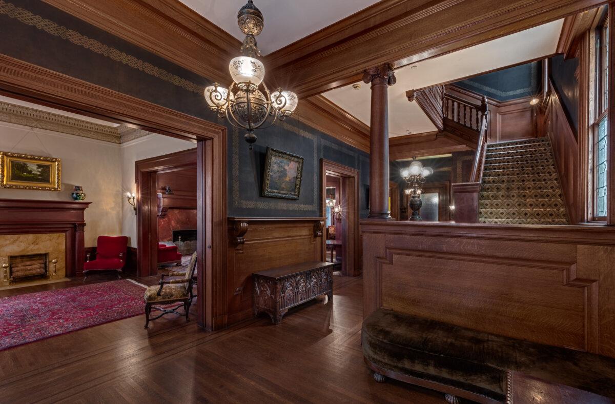 The first-floor entry boasts hardwood floors with an ornately carved hall table. The brass lighting fixture is authentic to the era as is the standing lamp. The stairs leading to the second floor are redwood as is the wainscoting, stair banisters, and newel posts. Leaded glass windows let in light at the bottom of the stairs, and an electric lamp is fixed on the stairway post. (Courtesy of Barry Schwartz Photography)