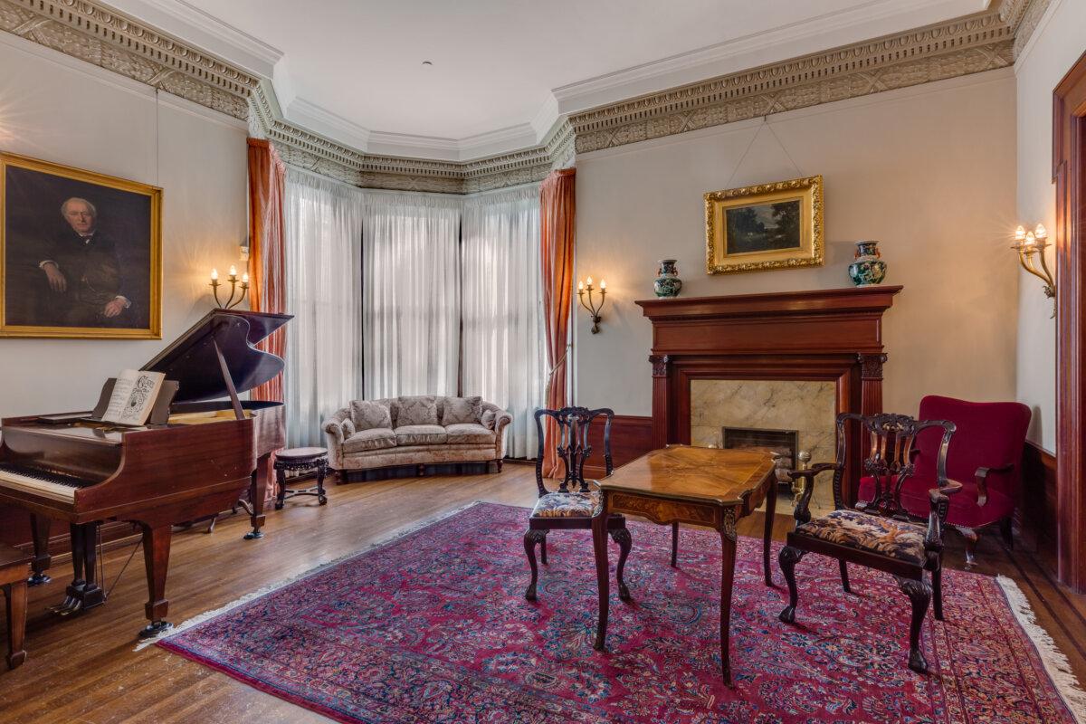 The Piano Room, or First Parlor, features a baby grand piano, shiny hardwood floors, and a Persian carpet. An intricately carved crown molding tops the walls. The sheer curtains sided by velvet tiebacks in the alcove are typical of the city due to its mild climate. The room is warmed by a marble fireplace with a mahogany mantle, and decorated with oriental vases popular at that time. The small inlaid table stands between two Queen Anne-style chairs, richly upholstered with handmade needlepoint fabrics. The room is lit with electric wall sconces and an unseen chandelier. (Courtesy of Barry Schwartz Photography)