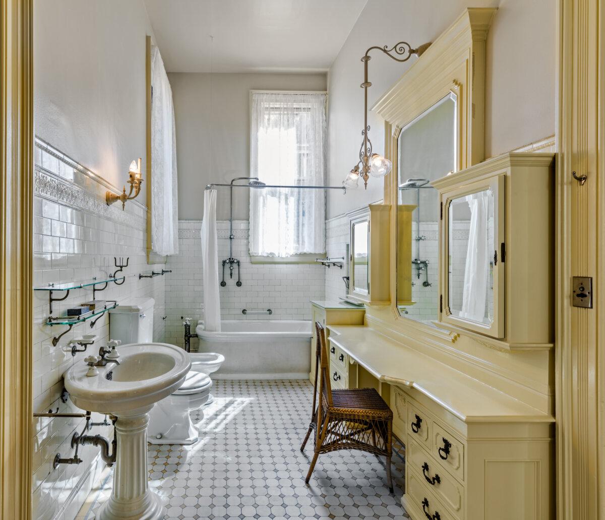 Although some of the fixtures have been updated in the bathroom, the vanity and cabinetry along the wall are original as is the tiled floor, tiled walls, and electric wall sconce lighting. Simple and white was traditional for family bathrooms of the day. A wicker chair sits along the soft yellow vanity and large mirror. (Courtesy of Barry Schwartz Photography)
