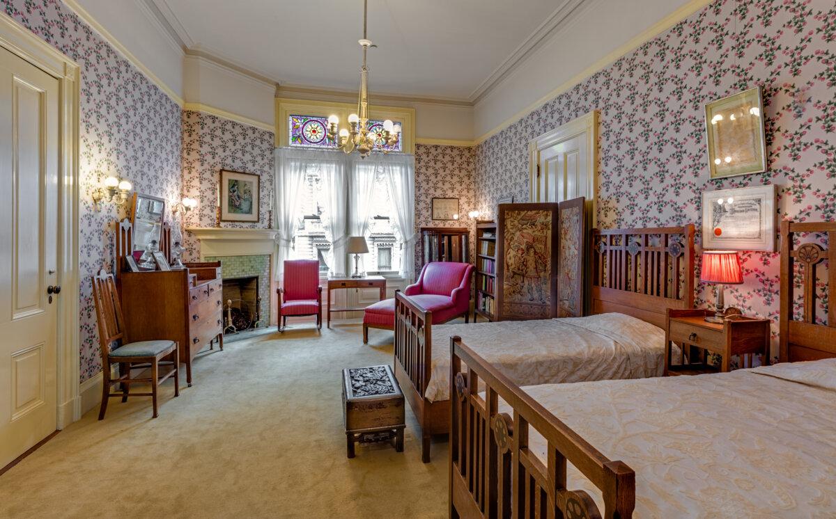 The back bedroom is on the second floor, with walls adorned with sizeable floral-patterned wallpaper and wall sconce lighting. A chandelier is in the center of the ceiling. The fireplace has been designed to scale, with a small mantle and a tiled front. The stained-glass windows above the plane glass windows with ruffled sheer curtains are typical of the Queen Anne style. A dressing screen next to a bed was used for modesty when changing, especially if servants were helping one to dress. (Courtesy of Barry Schwartz Photography)