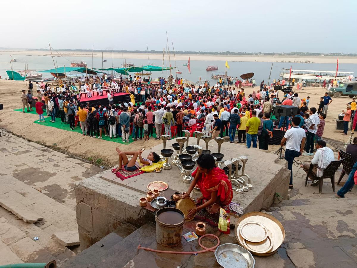 A woman washes temple wares at the Assi Ghat while a child surfs through her phone and a live election debate by Indian news channel Aj Tak takes place in the background, in Varanasi, Uttar Pradesh, India, on May 29, 2024. Varanasi is the constituency of Indian Prime Minister Narendra Modi. (Venus Upadhayaya/The Epoch Times)