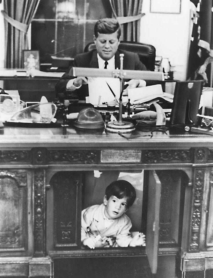 John Kennedy Jr. peeks out through the kneehole panel of the Resolute desk while his father, President John F. Kennedy, works. (Public Domain)