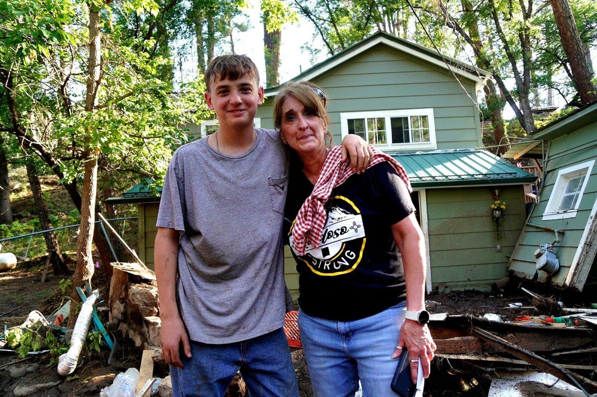 Stephanie Sheffield (R) and her grandson, Matthew, stand in front of her house, left filled with mud following flash flooding through the village of Ruidoso, N.M., on July 8, 2025. (Allan Stein/The Epoch Times)