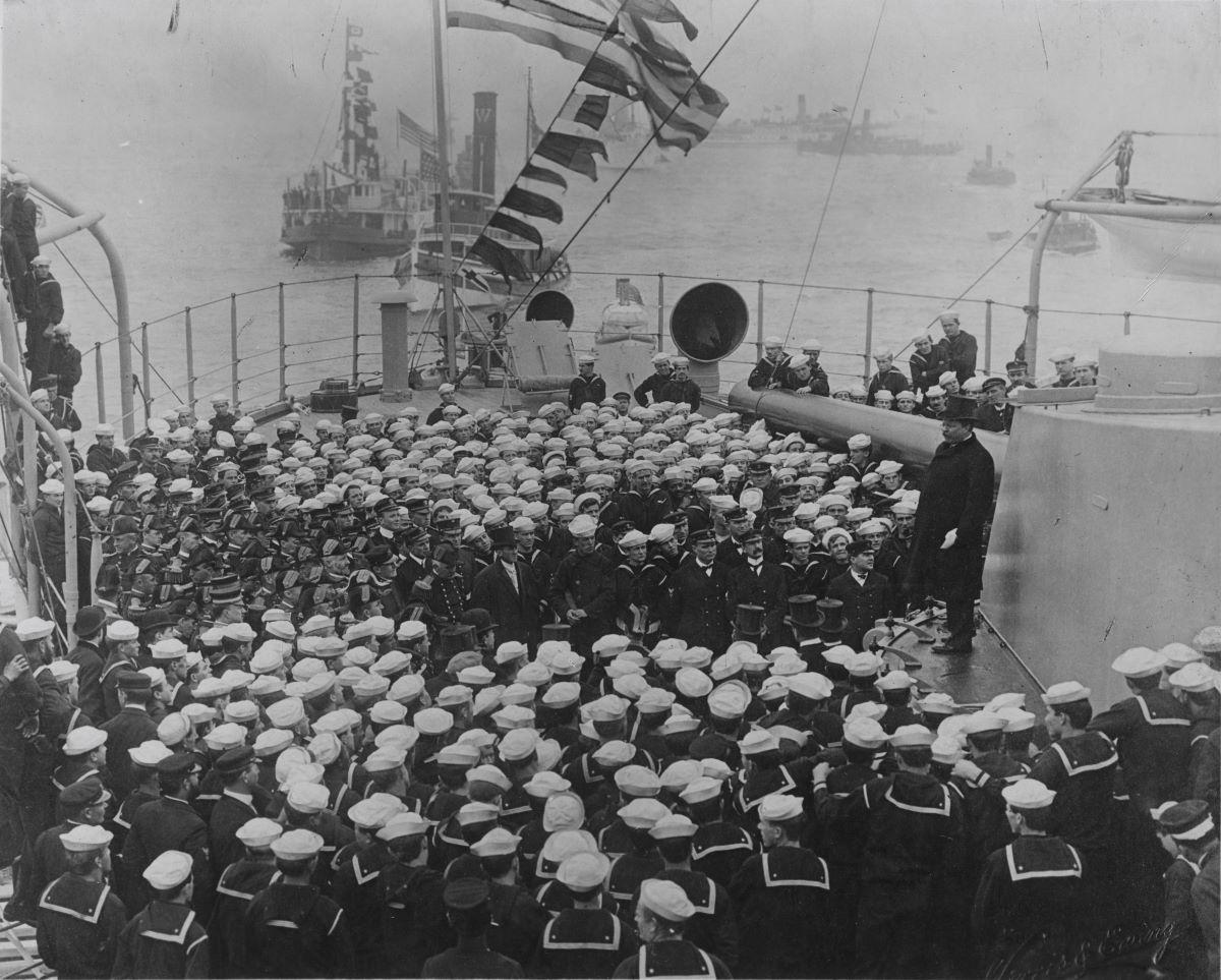 President Theodore Roosevelt, (on gun turret, R) addressing officers and crew on Connecticut, in Hampton Roads, Va., Feb. 22, 1909, upon the ship's return after circumnavigating the globe. (Public Domain)