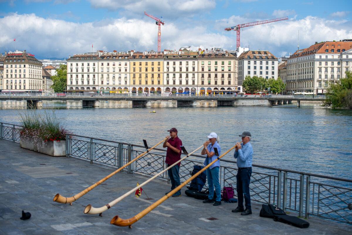 Buildings topped with the logos of Vacheron Constantin, Zenith, Baume & Mercier, and Breitling are visible across Lake Geneva, with horn players performing in the foreground on the opposite shore in Geneva, Switzerland, on Aug. 2, 2025. (Robert Hradil/Getty Images)