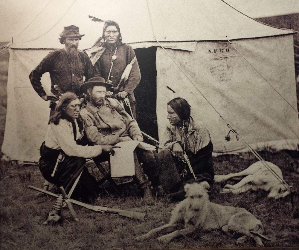 The Indian Scout Bloody Knife (L) kneels next to Custer (center). Staghounds lounge in the foreground. (Public Domain)
