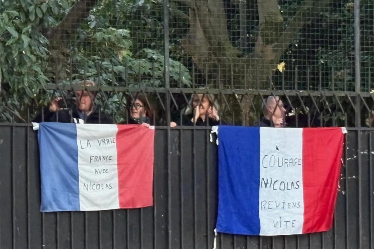 People stand behind French flags with inscriptions reading "True France with Nicolas" at left and "Courage Nicolas, come back soon" outside former French President Nicolas Sarkozy's home in Paris on Oct. 21, 2025. (Masha Macpherson/AP Photo)