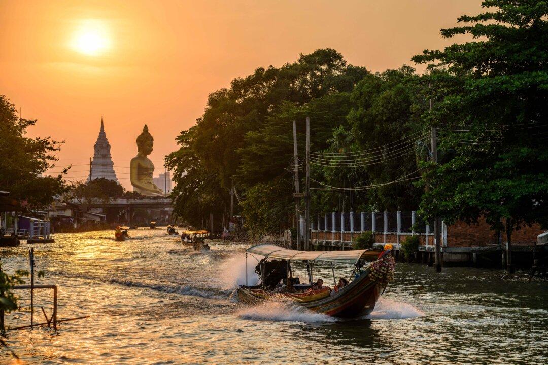 People ride boats in a canal as the sun begins to set behind the 69-metre tall giant Buddha statue of Wat Paknam Phasi Charoen temple (back, L) in Bangkok on February 16, 2026. (Photo by Anthony WALLACE / AFP via Getty Images)