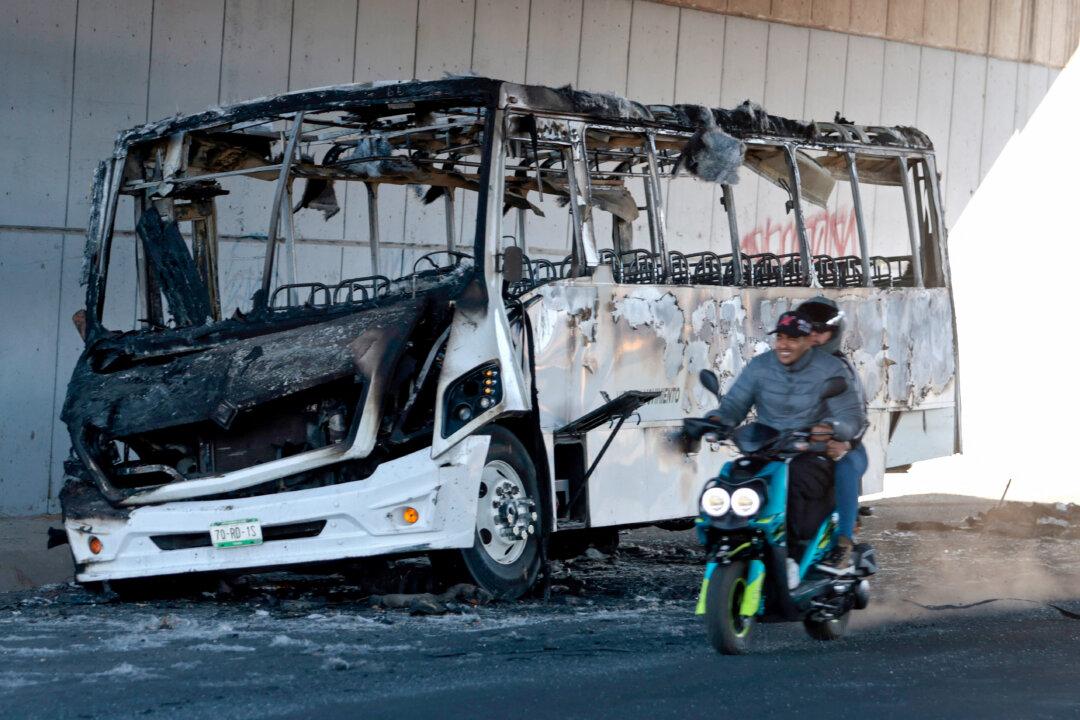 A motorcycle rides past a burned bus used to block an avenue, is seen in Tonala, Jalisco, Mexico, on February 23, 2026. Mexico has deployed 10,000 troops to quell clashes sparked by the killing of the country's most-wanted drug lord, Nemesio "El Mencho" Oseguera, leader of the Jalisco New Generation Cartel (CJNG), that have claimed dozens of lives, officials said on February 23. News of his death triggered spasms of violence, with cartel members blocking roads in 20 states and torching vehicles and businesses. (Photo by Ulises RUIZ / AFP via Getty Images)