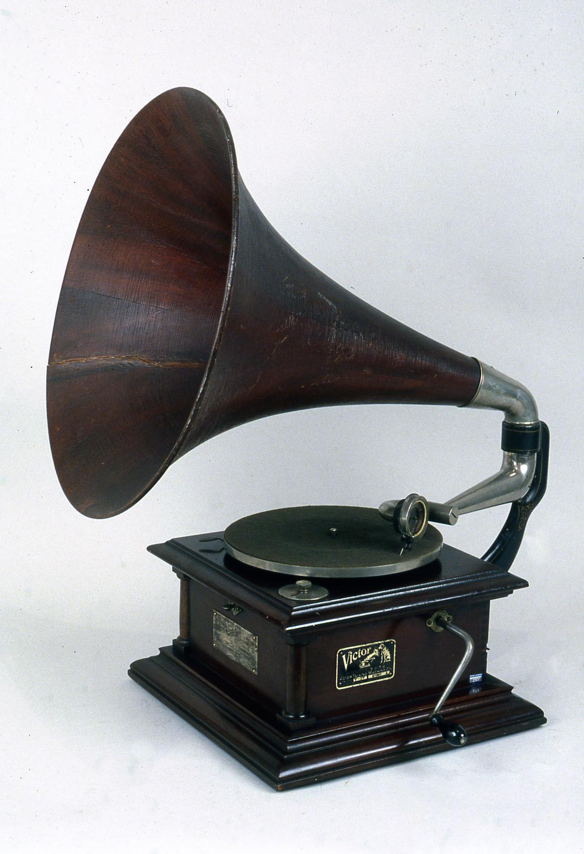 A detail view of a 1906 gramophone with a mahogany cabinet that houses the turntable and a horn. A double spring-driven motor is operated by a crank located on the right side of the base. National Museum of Science and Technology, Milan, Italy. (<a class="external text" href="http://www.museoscienza.org/dipartimenti/catalogo_collezioni/scheda_oggetto.asp?idk_in=ST120-00150" rel="nofollow">Catalogo collezioni</a>/<a title="Category:CC-BY-SA-4.0" href="https://commons.wikimedia.org/wiki/Category:CC-BY-SA-4.0">CC-BY-SA-4.0</a>)