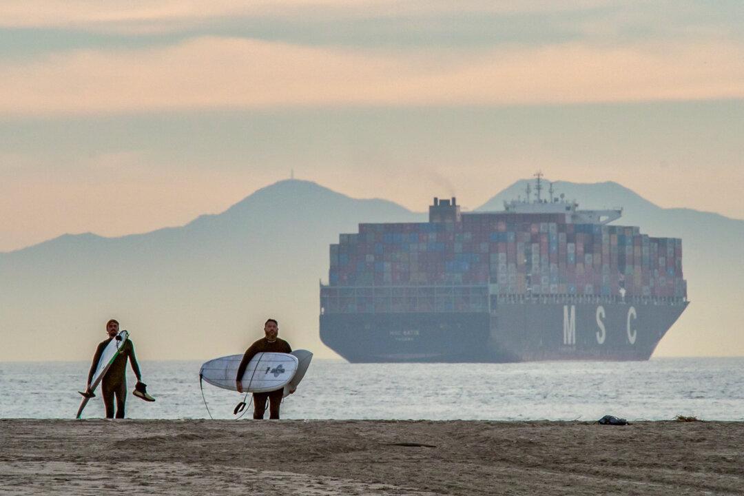 Surfers leave the water as a vessel laden with shipping containers looms off the coast of Seal Beach, Calif., on Jan. 12, 2021. Most manufactured goods traded internationally are transported in these “boxes,” which have become the units of global trade. (John Fredricks/The Epoch Times)