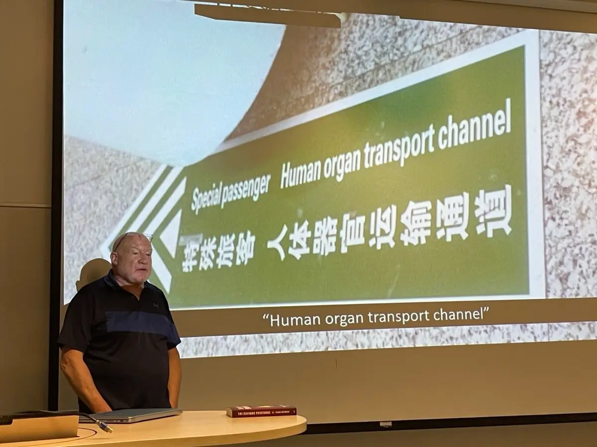 Author Ethan Gutmann stands next to a photo of a sign indicating a fast processing lane for organs at the Aksu airport in China, during a panel of the International Congress on Law and Mental Health in Montreal on July 1, 2026. (Noé Chartier/The Epoch Times)