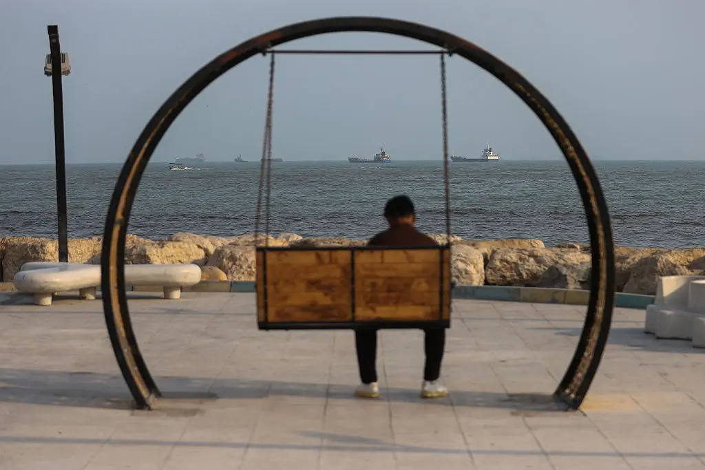 A person sits on a swing along the shoreline as ships are seen anchored in the Strait of Hormuz off the coast of Bandar Abbas, Iran, on August 10, 2026. (Ali Saeedi/Getty Images)