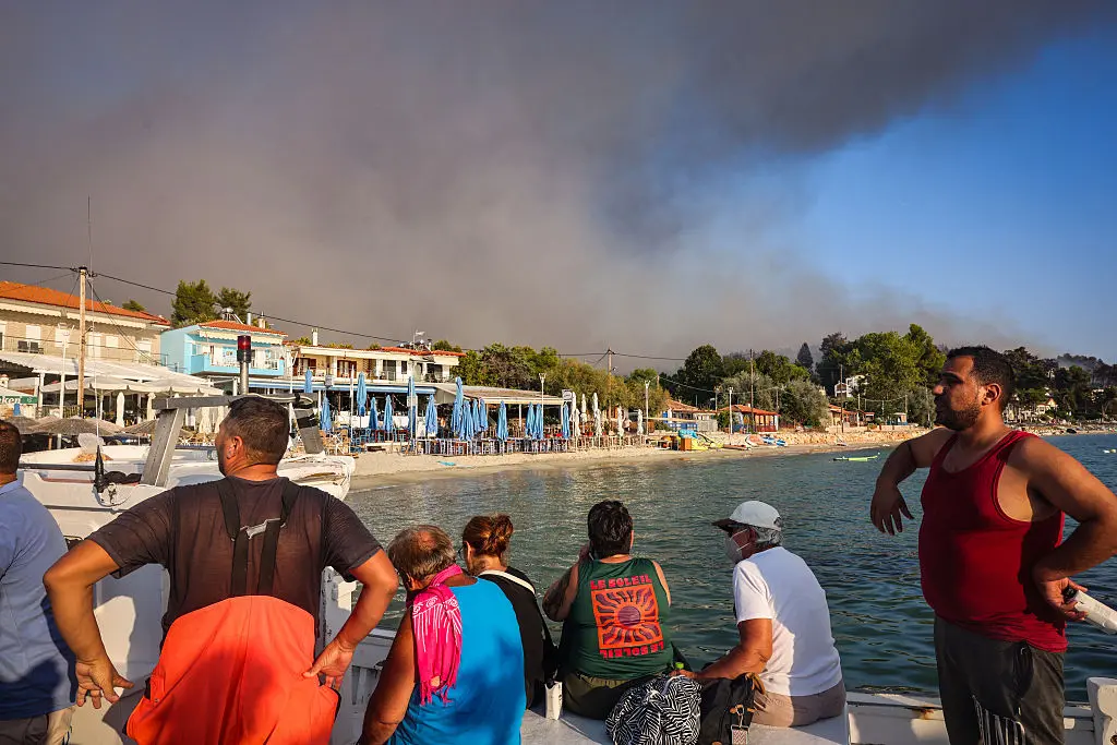 People evacuate on small boats during a wildfire as a plume of smoke rises into the sky near the village of Siviri, northern Greece, on Aug. 13, 2026. A fleet of small boats evacuated hundreds of tourists from beaches in northern Greece as a forest fire bore down on two popular resorts. (Vasilis Ververidis/AFP via Getty Images)