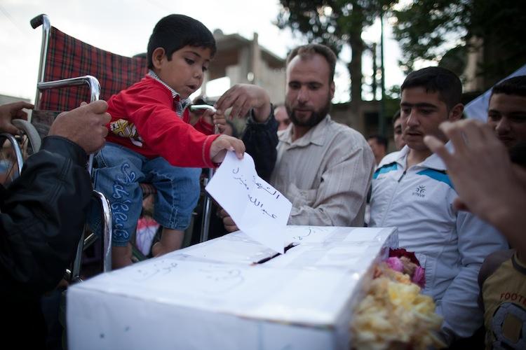 <a><img class="size-large wp-image-1787796" title="A handicapped Syrian boy casts a paper bearing the name of a 'martyr' into a coffin-like symbolic ballot box during an anti-election demonstration in the city Qusayr, Syria, on May 7. (STR/AFP/GettyImages)" src="https://www.theepochtimes.com/assets/uploads/2015/09/SyriaElections143990501.jpg" alt="" width="590" height="393"/></a>
