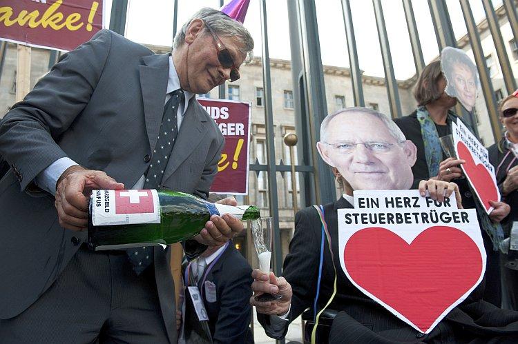 <a><img class="size-large wp-image-1788995" title="A demonstrator carrying a bottle of 'Schwarzgeld' (dirty money) champagne" src="https://www.theepochtimes.com/assets/uploads/2015/09/bottle125712977.jpg" alt="A demonstrator carrying a bottle of 'Schwarzgeld' (dirty money) champagne" width="590" height="392"/></a>
