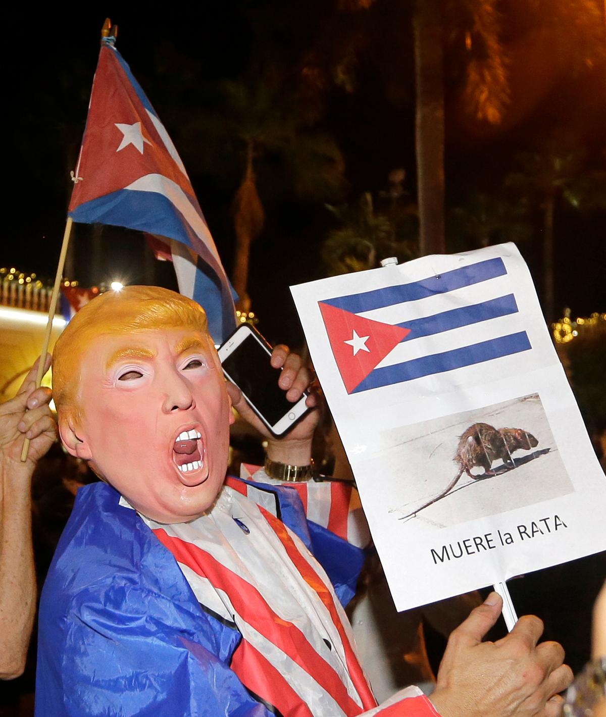 A person wearing a Donald Trump mask holds a sign in Spanish that reads the Rat Dies as he celebrates the death of Fidel Castro in the Little Havana area in Miami., on Nov. 26, 2016. (AP Photo/Alan Diaz)
