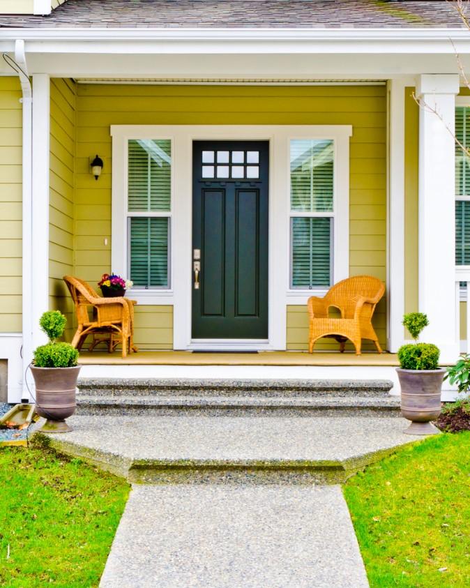 Natural wicker chairs and an ochre-painted house with white trim create a welcoming entryway. The sculptured topiaries flanking the front door add a nice touch. (Karamysh stock photo/Shutterstock)