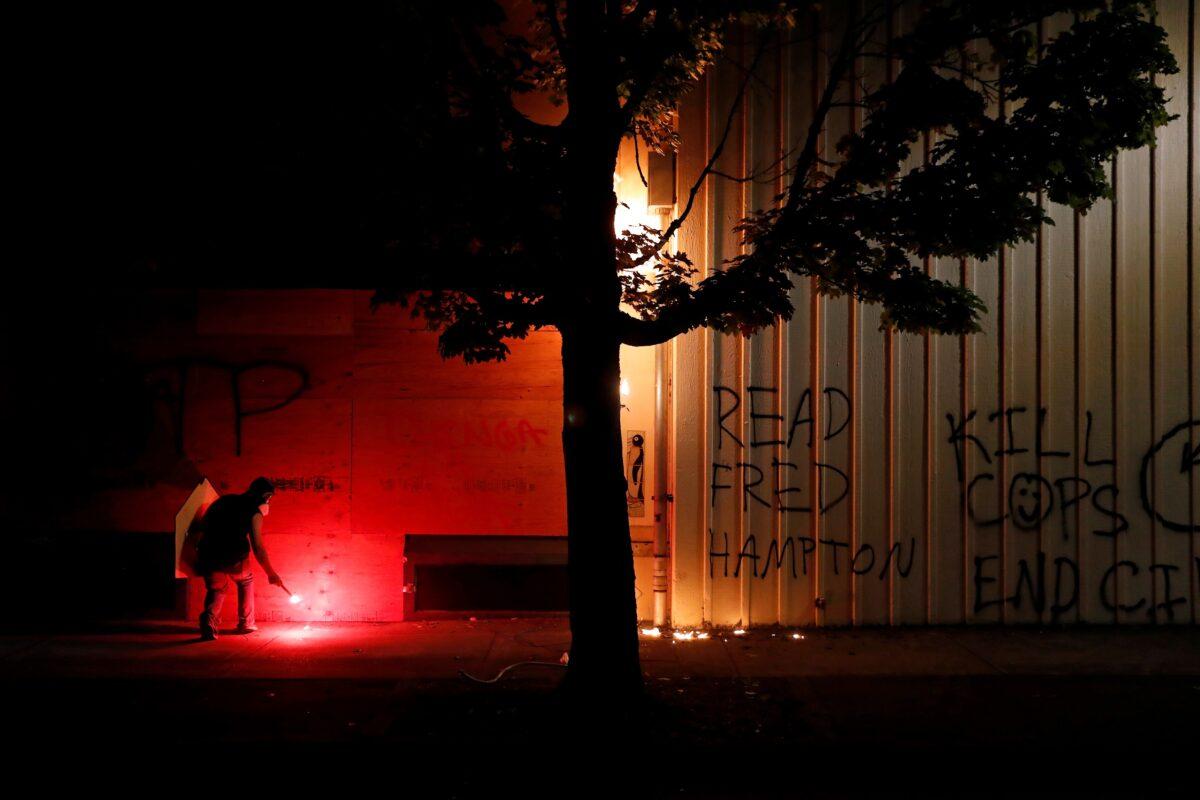 A demonstrator picks up a lit flare while a fire burns on the Portland Police North Precinct during a riot in Portland, Ore., Aug. 23, 2020. (Terray Sylvester/Reuters)
