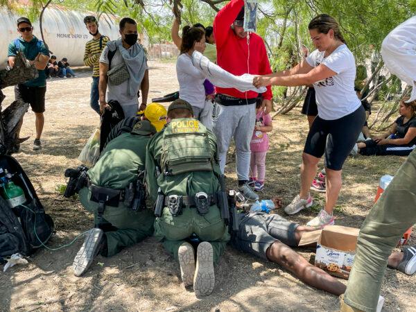 Border Patrol agents provide medical assistance to an illegal immigrant who has heat-related issues, while other agents apprehend a large group of illegal immigrants near Eagle Pass, Texas, on May 20, 2022. (Charlotte Cuthbertson/The Epoch Times)